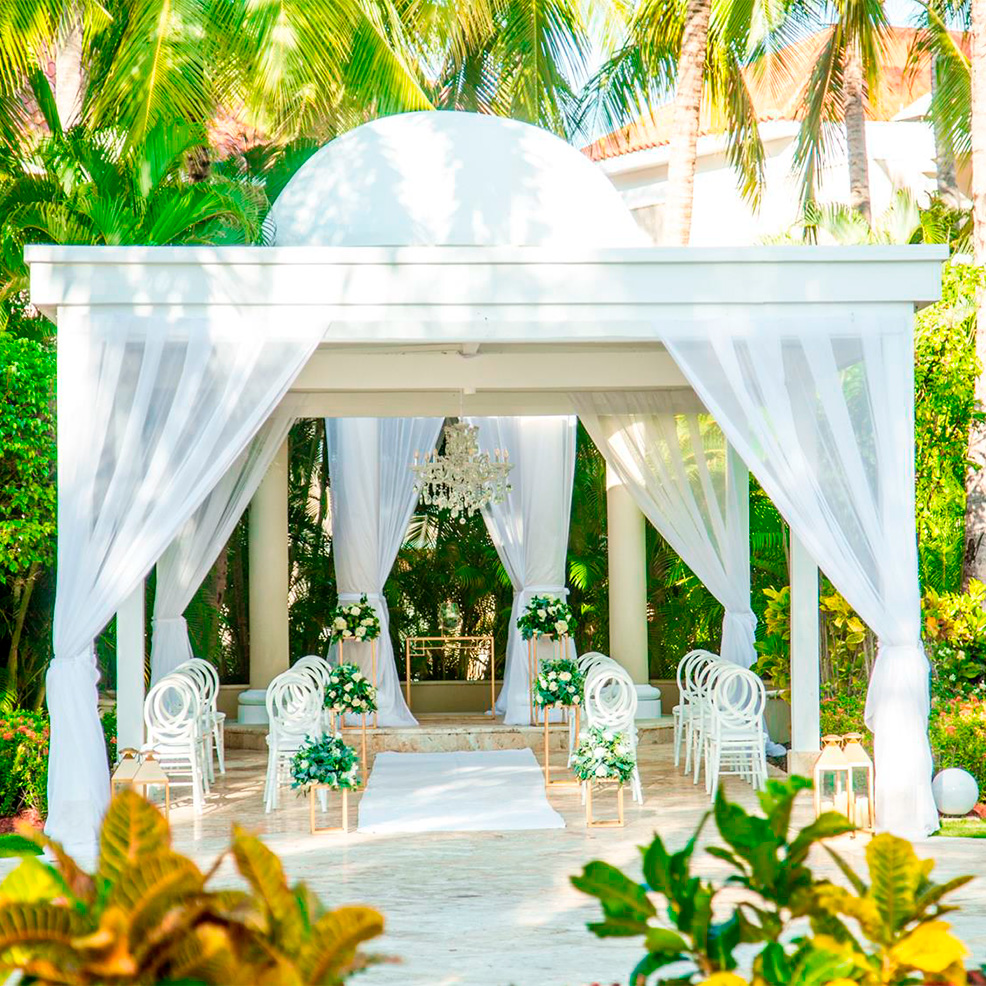 An outdoor wedding venue features a white gazebo with sheer drapes, white chairs, and floral arrangements. Palm trees and greenery surround the setting, creating a tropical ambiance.