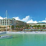 A tropical resort destination for weddings, featuring a large hotel building, palm trees, a sandy beach with loungers, and a sailboat on clear turquoise water under a blue sky.