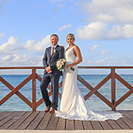 A bride and groom smiling, standing on a wooden pier by the sea at their destination wedding, with the groom sitting on a railing and the bride holding a bouquet.