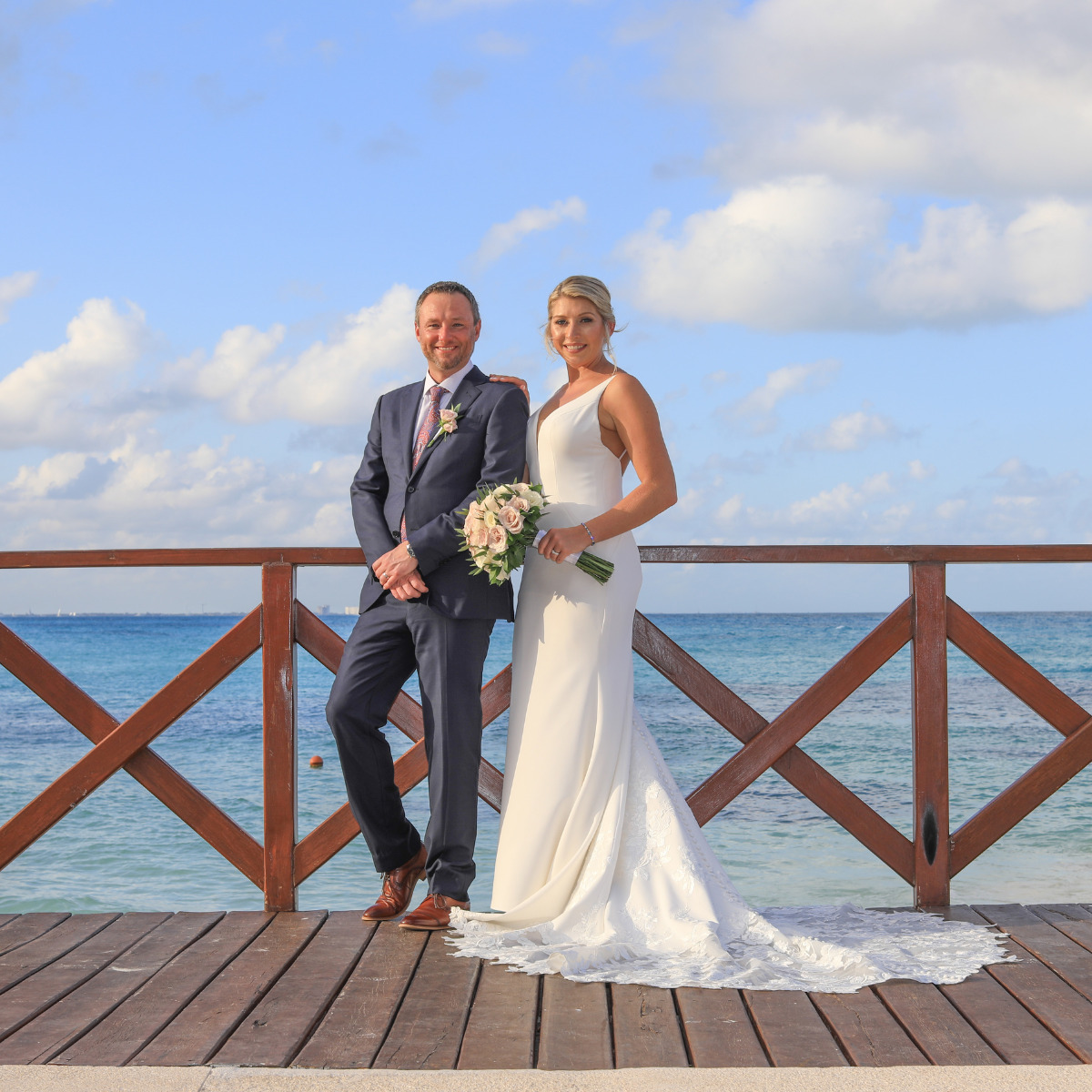 A bride and groom smiling, standing on a wooden pier by the sea at their destination wedding, with the groom sitting on a railing and the bride holding a bouquet.