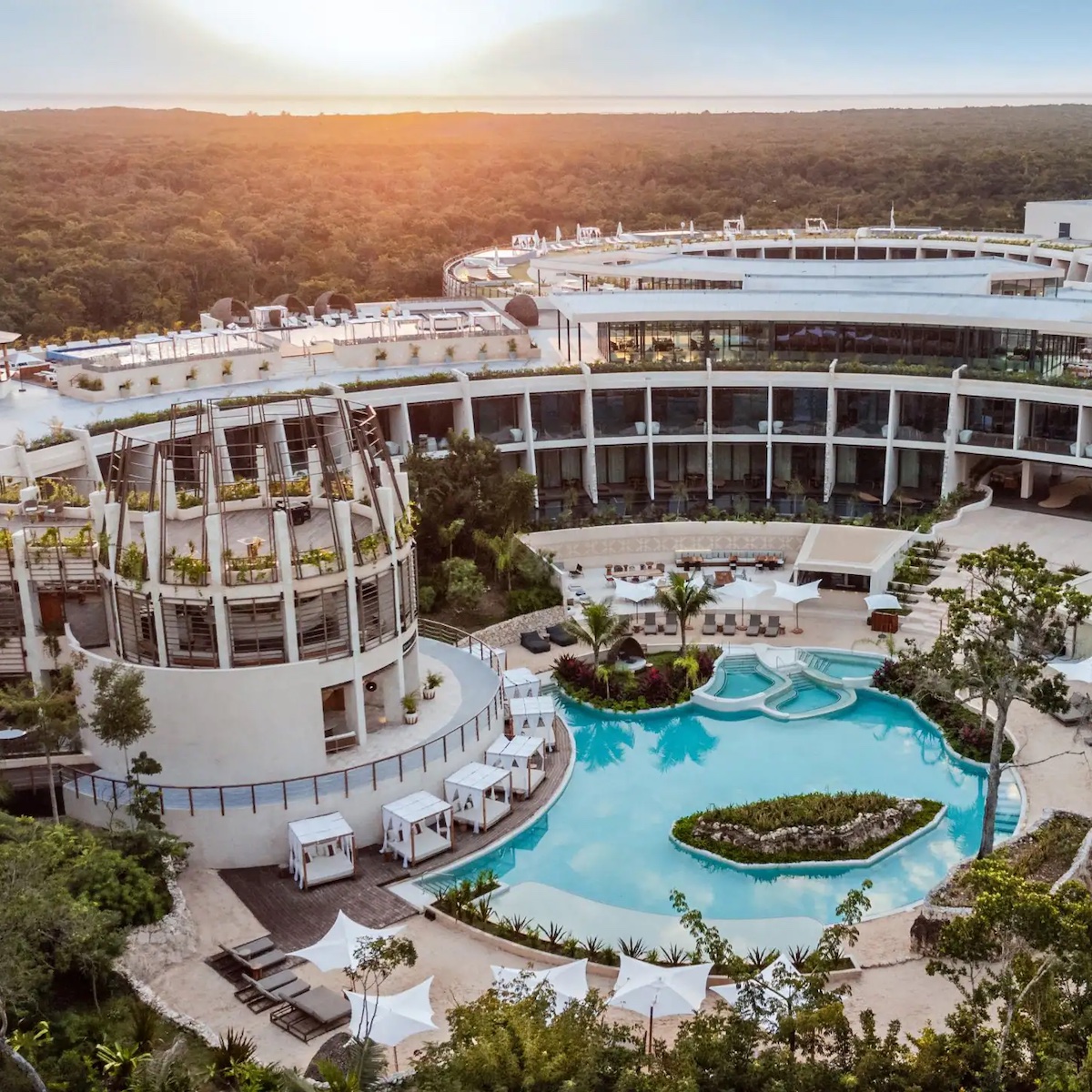 Aerial view of Secrets Tulum, showcasing a circular building and a pool embraced by lush greenery under a setting sun.