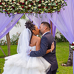 A bride and groom kissing under a floral arch with purple drapery at a destination wedding ceremony.