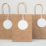 Three brown paper bags with handles, ideal for wedding gift ideas, each featuring a large white circular tag, standing on a tabletop against a white background.