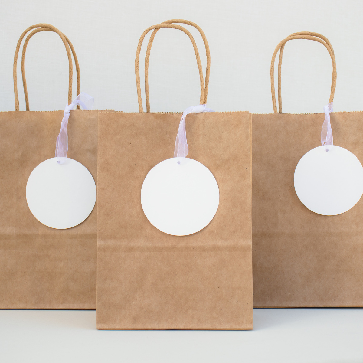 Three brown paper bags with handles, ideal for wedding gift ideas, each featuring a large white circular tag, standing on a tabletop against a white background.