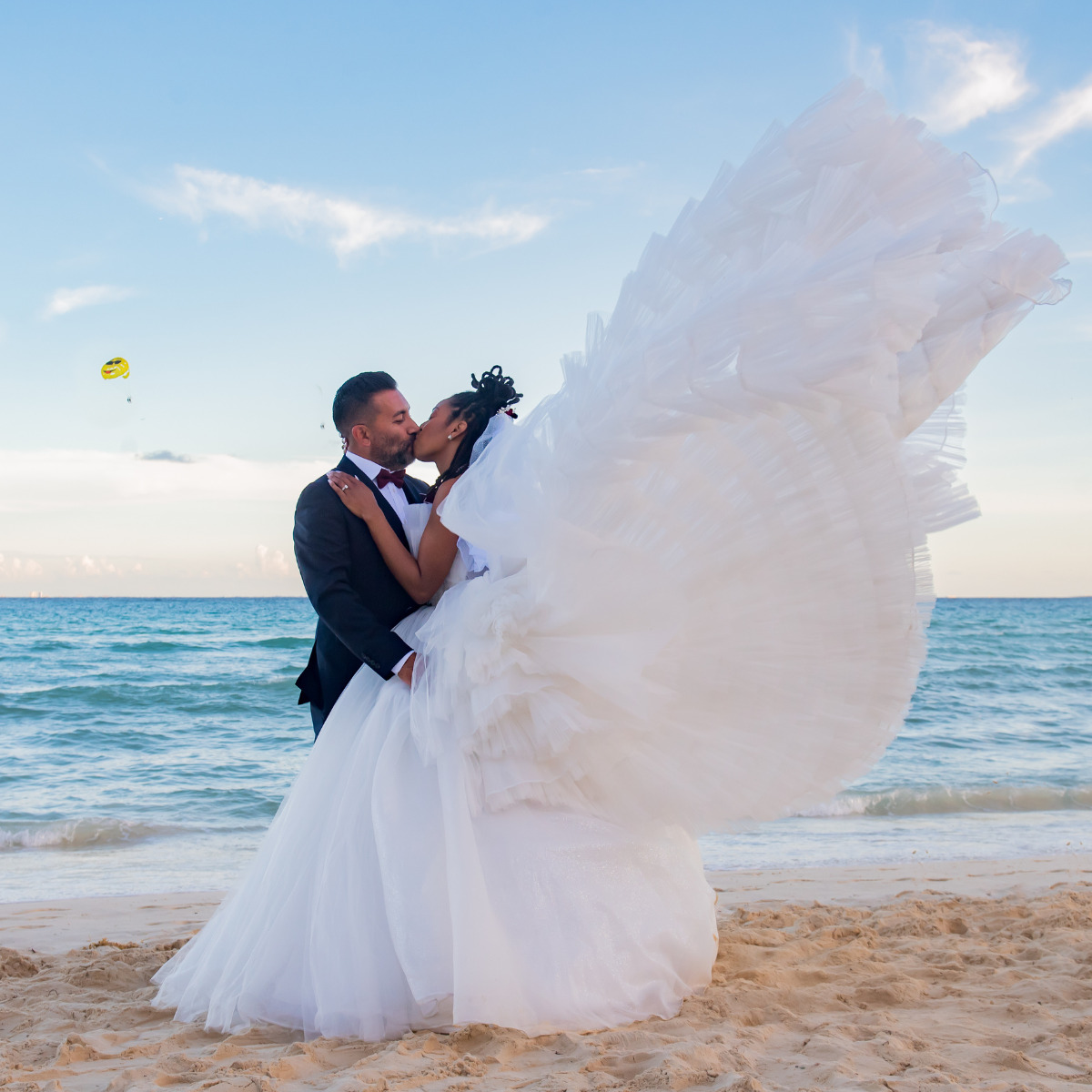 A Aushiree and Robert in wedding attire embracing on a destination wedding beach with the bride's dress trailing in the wind.