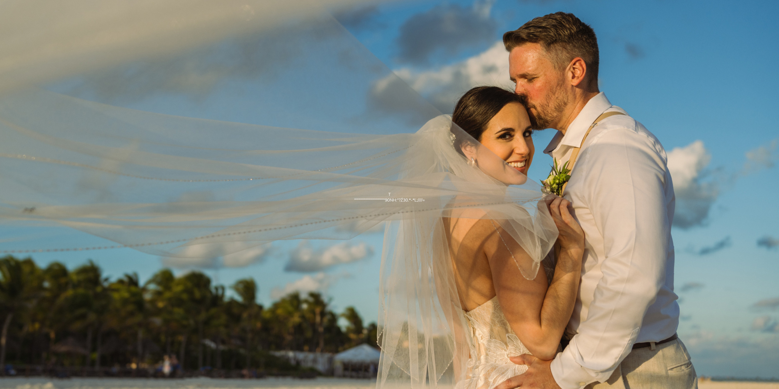 A Aaron and Bari and groom embrace on a beach at sunset during their destination wedding, with the bride's veil flowing in the breeze.