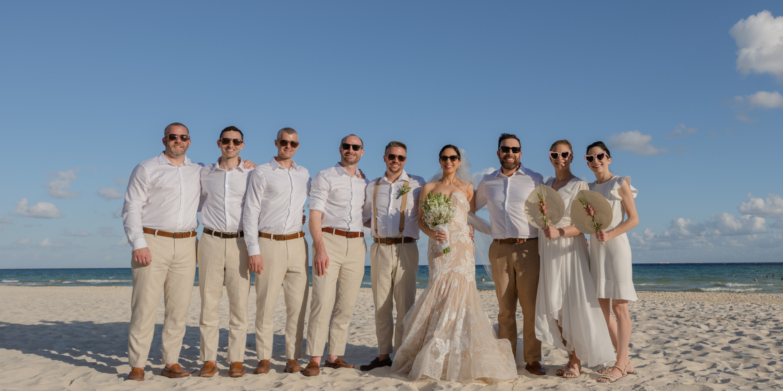 A bride and groom with eight guests dressed in white shirts and beige pants stand on a beach under a clear blue sky at their Aaron and Bari destination wedding.