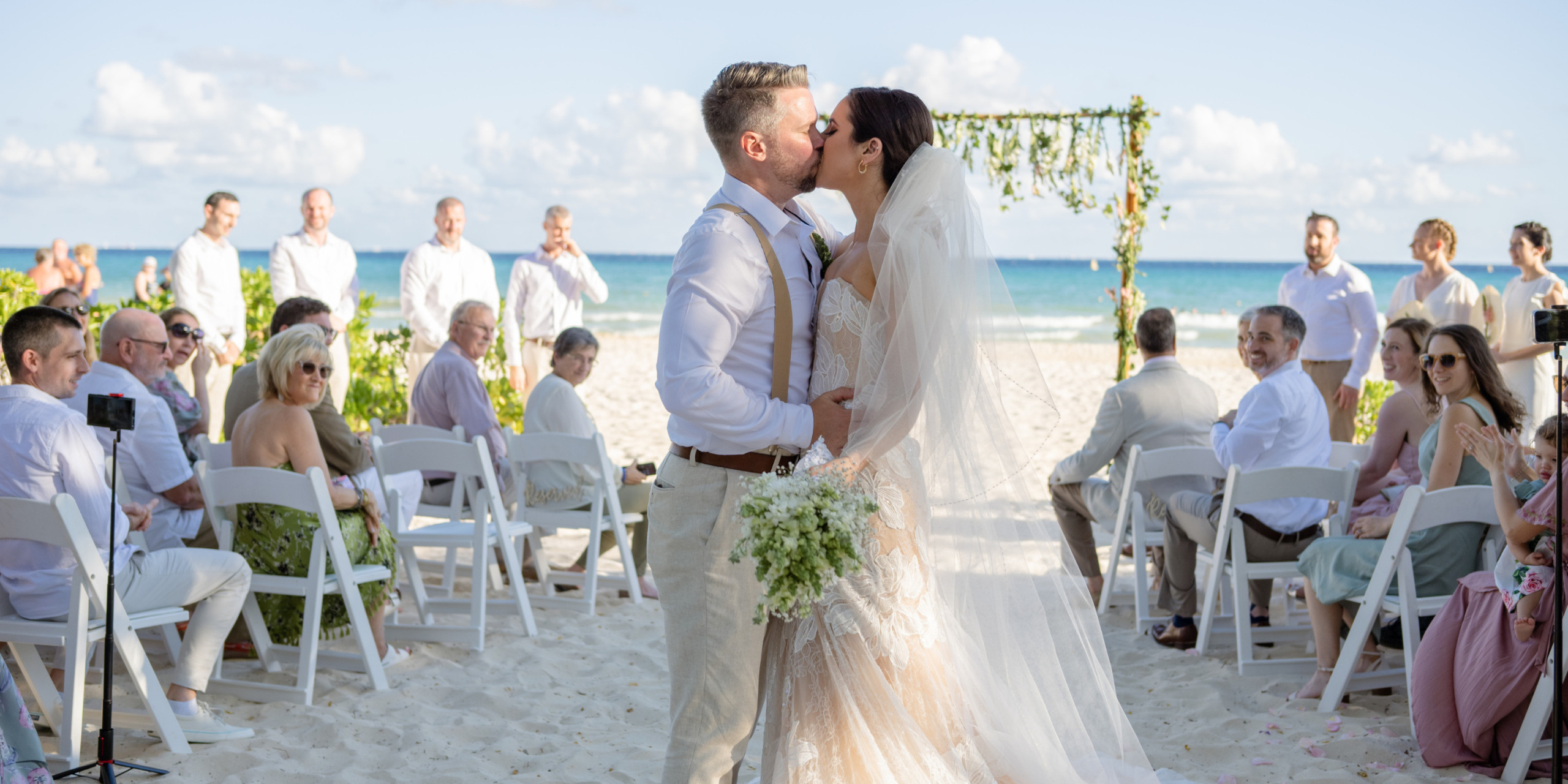 A couple kissing at their Aaron and Bari wedding ceremony on the beach with guests seated around them under a clear sky.