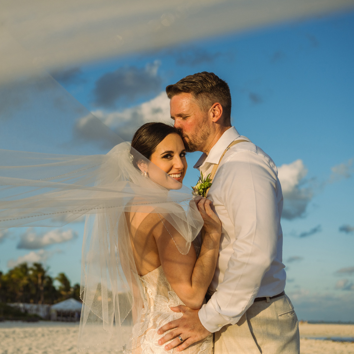 Aaron and Bari embracing on a beach at sunset during their destination wedding, with the bride's veil blowing in the wind.