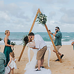 A couple kisses under a wooden arch on a beach at their Destify wedding ceremony, with guests, including a child, watching.