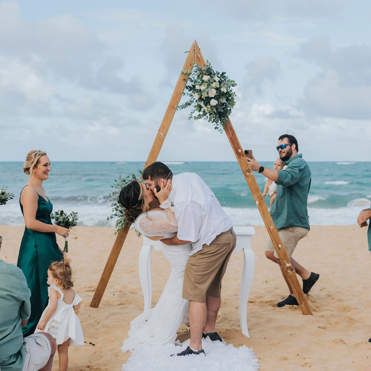A couple kisses under a wooden arch on a beach at their Destify wedding ceremony, with guests, including a child, watching.