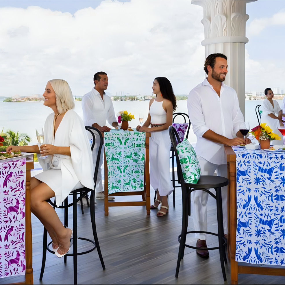 Four people standing by patterned high tables at a waterfront bar, casually dressed and smiling, with tropical drinks and flowers on tables during a destination wedding.