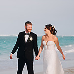 A bride in a white dress and a groom in a black tuxedo holding hands on a sandy beach at their international destination wedding, smiling as they walk.