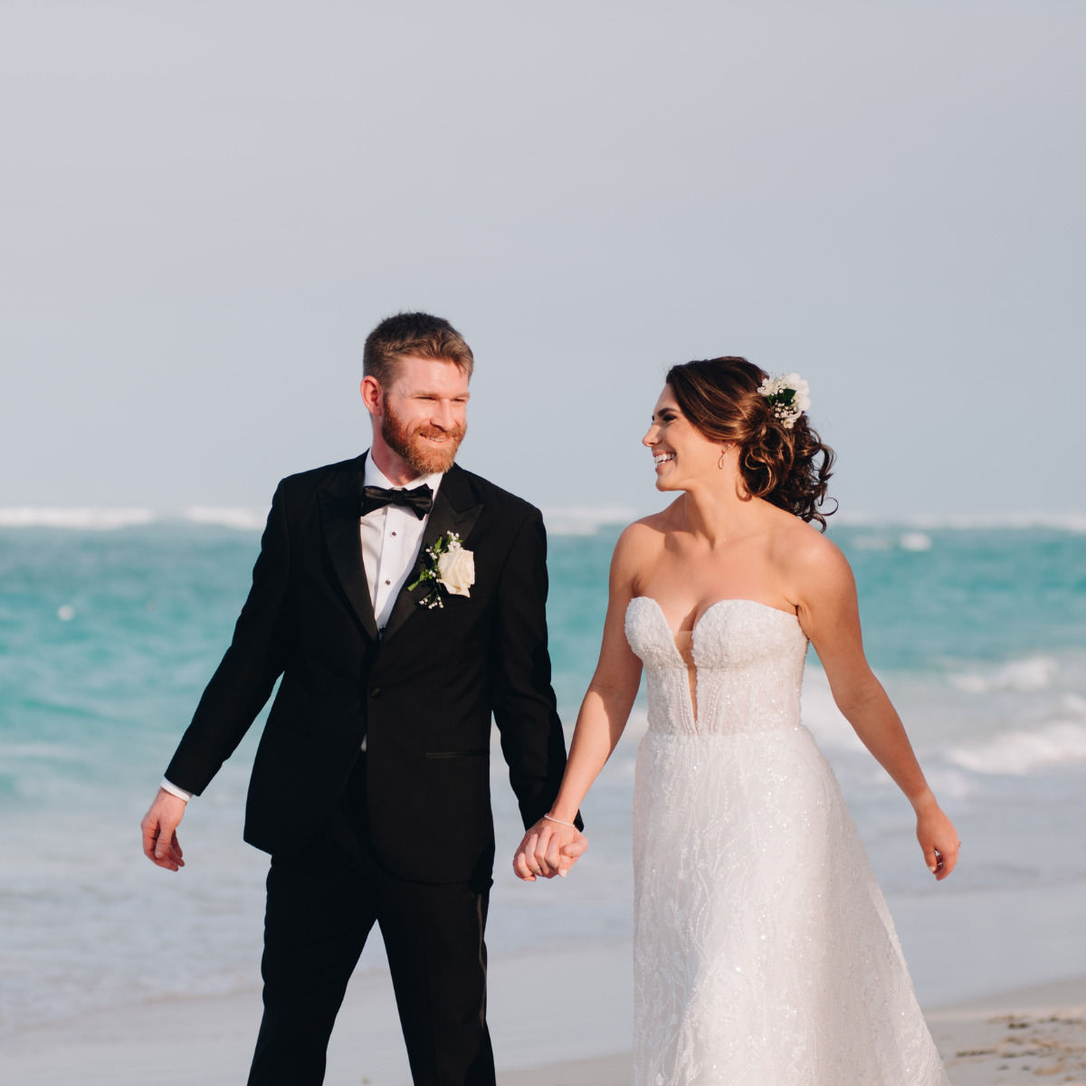 A bride in a white dress and a groom in a black tuxedo holding hands on a sandy beach at their international destination wedding, smiling as they walk.
