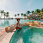A woman in a white wedding swimsuit smiles while sitting on rocks at the edge of a resort pool, with a palm-thatched umbrella and clear skies in the background.