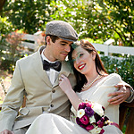 A bride and groom smiling and sitting closely on a bench outdoors at their destination wedding, the bride holding a bouquet of purple flowers.