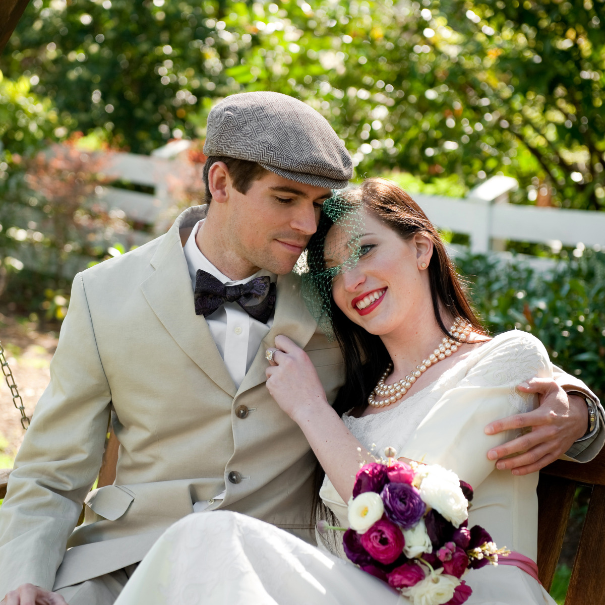 A bride and groom smiling and sitting closely on a bench outdoors at their destination wedding, the bride holding a bouquet of purple flowers.