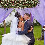 A bride and groom kissing under a floral arch with purple drapery at their wedding ceremony.