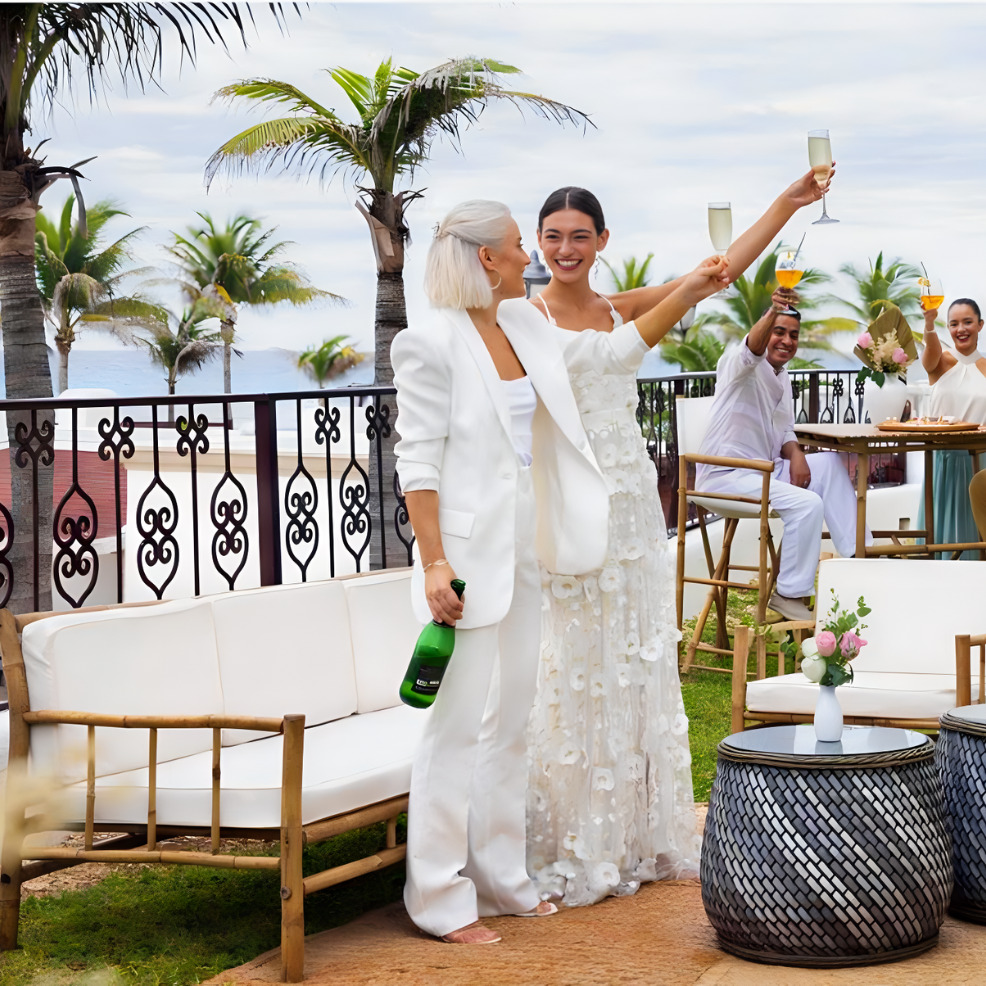 Two women in elegant white outfits celebrating with champagne at a destination wedding, with guests clapping in the background.