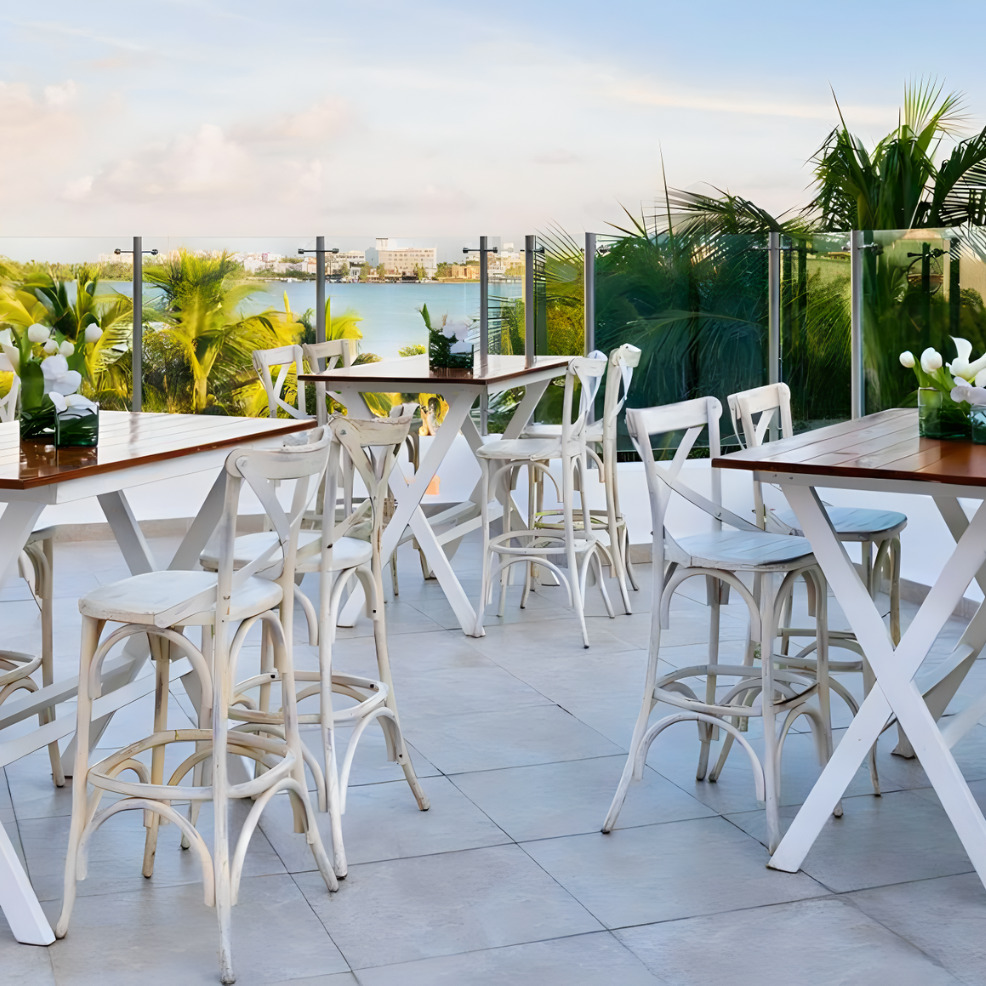 Outdoor dining area with white tables and chairs on a terrace overlooking a scenic waterfront at sunset, perfect for a destination wedding.