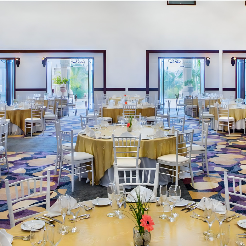 Elegant dining hall set for a destination wedding with round tables, white chairs, and floral centerpieces, under a bright, airy ambiance.
