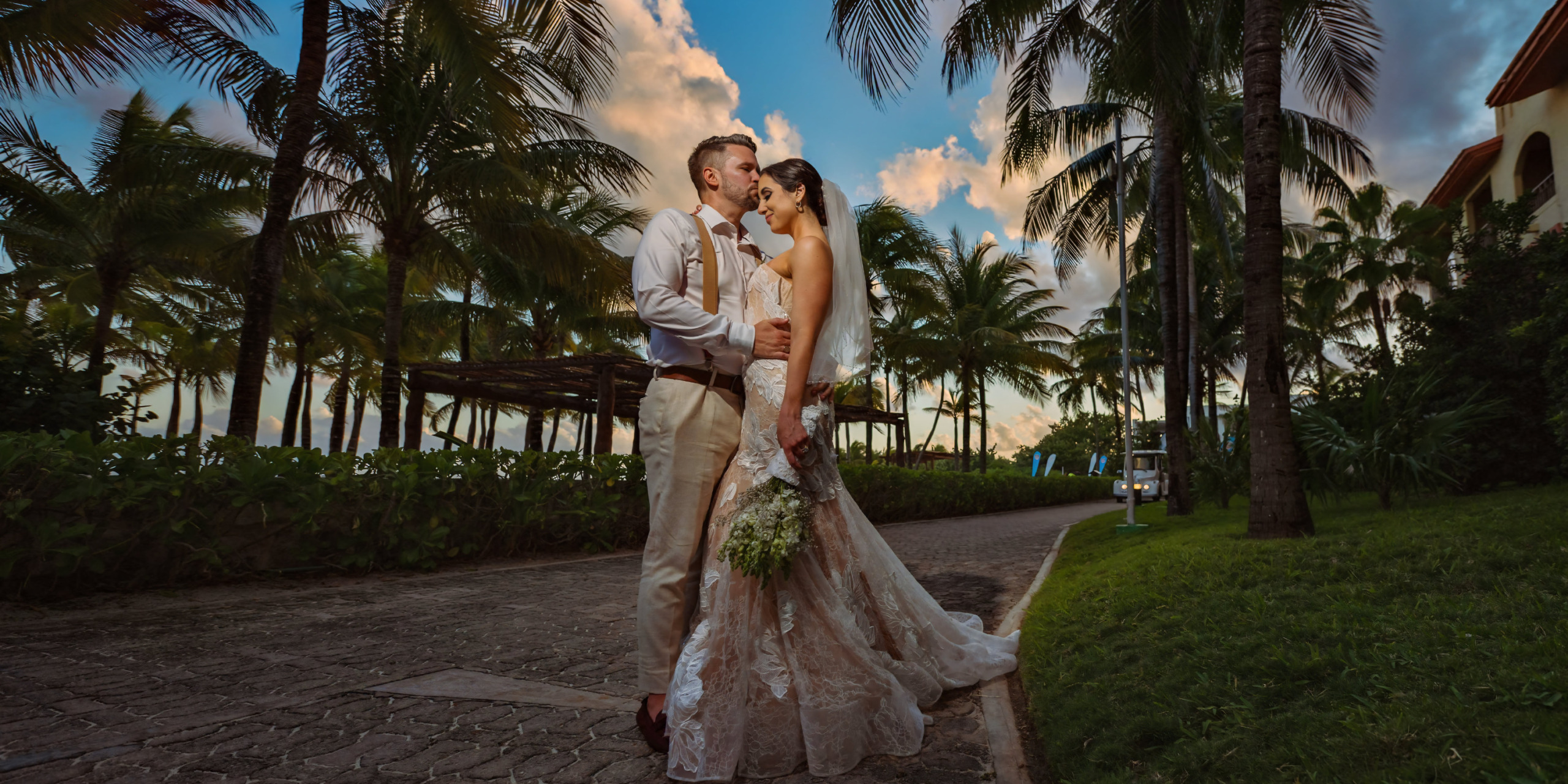 A Aaron and Bari kiss in front of palm trees at sunset during their destination wedding.