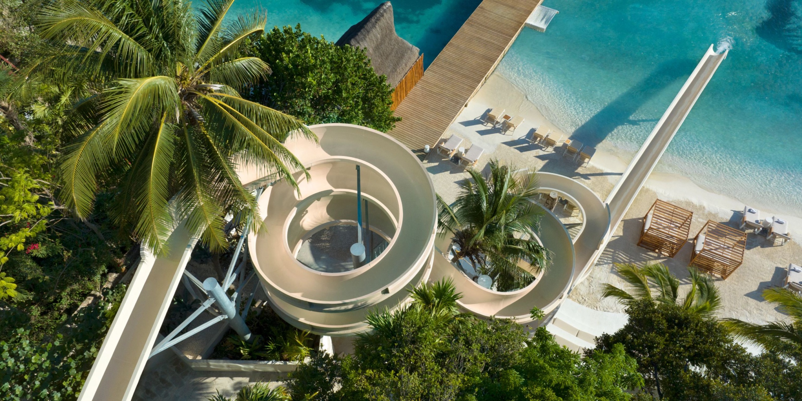 Aerial view of the water slide at Impression Isla Mujeres by Secrets, winding around palm trees and leading to a blue pool near a sandy beach adorned with lounge chairs and thatched umbrellas.