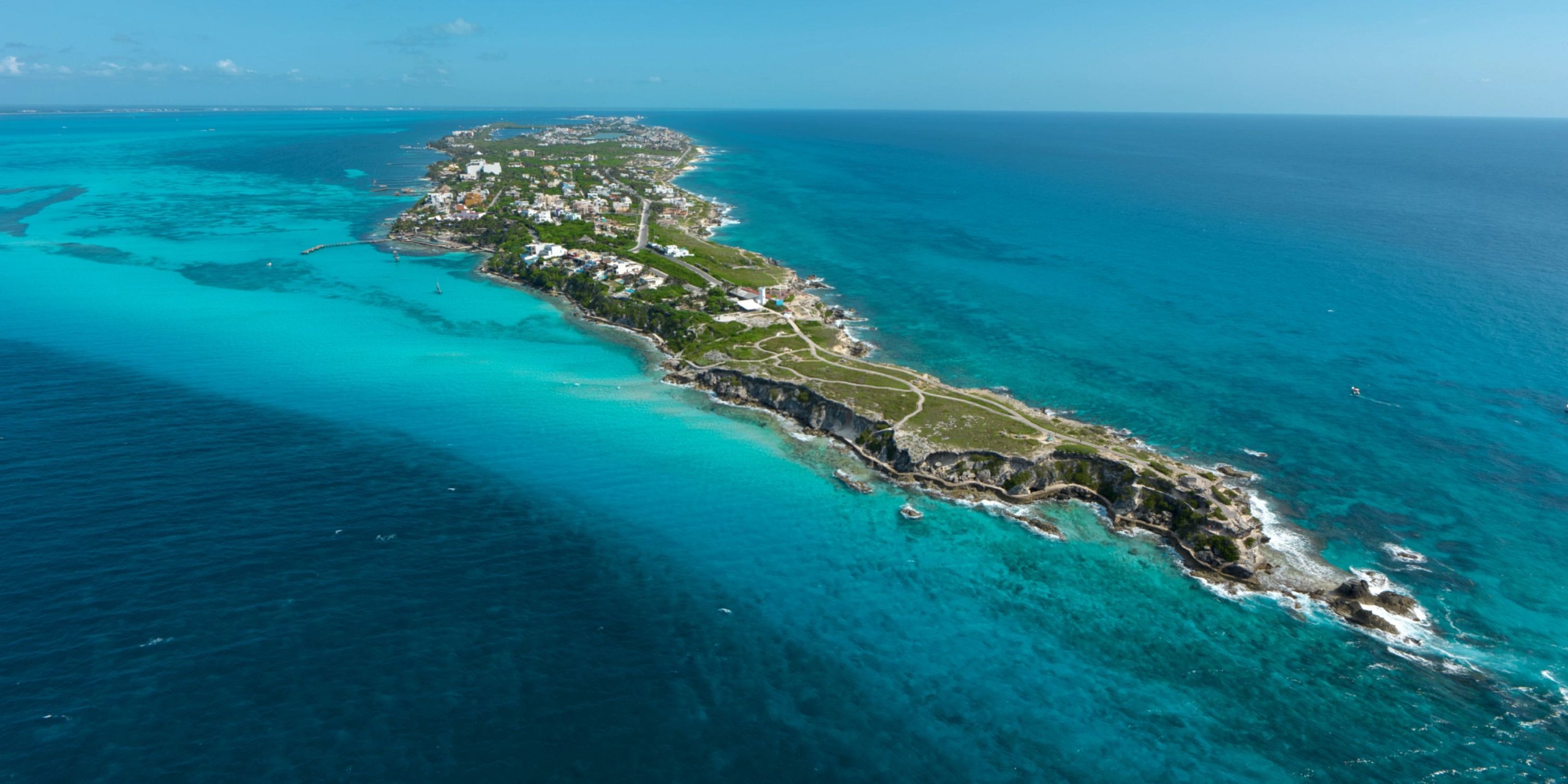 Aerial view of Impression Isla Mujeres by Secrets, a narrow island encircled by turquoise and deep blue ocean waters, featuring lush green vegetation and scattered buildings along its coastline.
