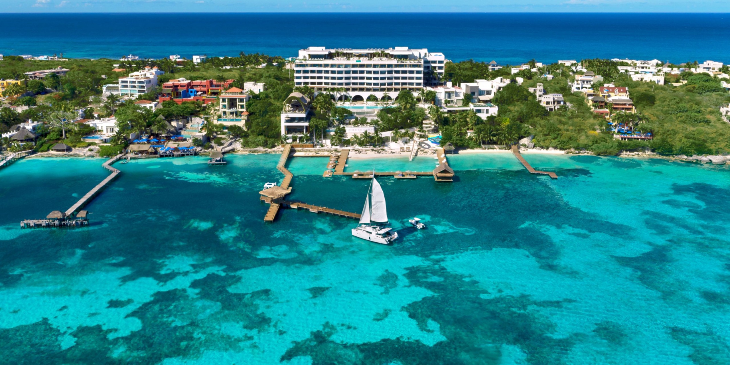 An aerial view of Impression Isla Mujeres by Secrets showcases a coastal resort with pristine white buildings, encircled by crystalline turquoise waters. A sailboat rests docked alongside wooden piers that stretch into the bay, while lush greenery frames the scenic area.