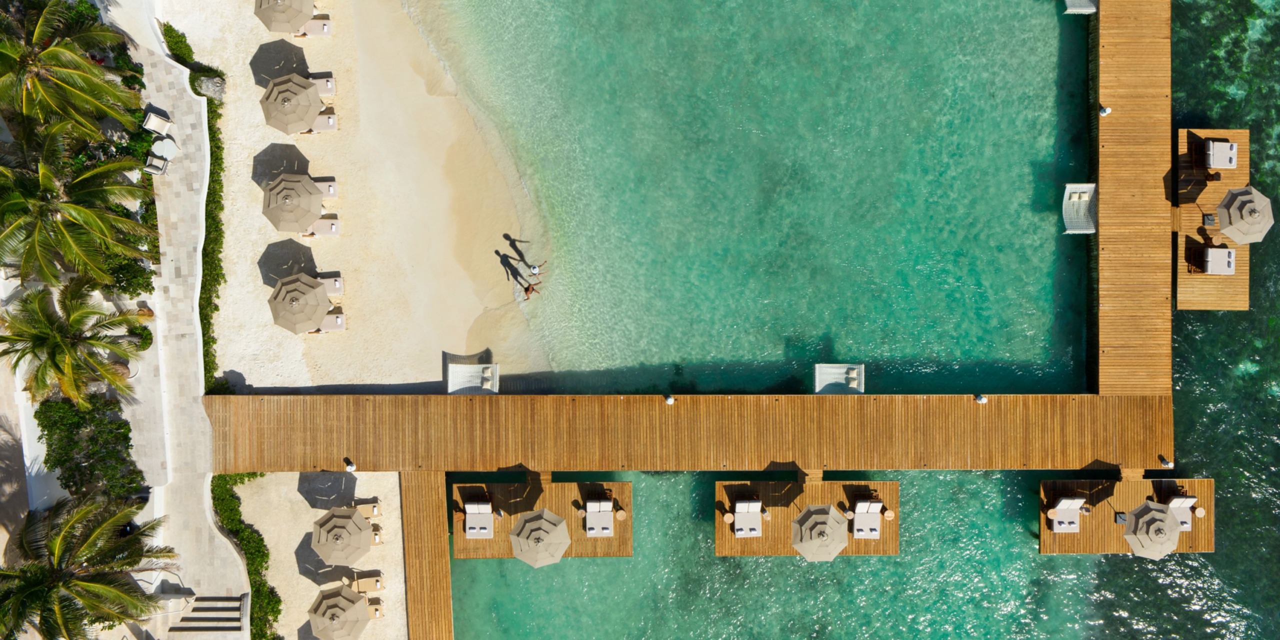 Aerial view of Impression Isla Mujeres by Secrets, featuring a beach with a wooden pier, sun loungers, and umbrellas both on the sand and over the turquoise water. Two people stroll along the shoreline.