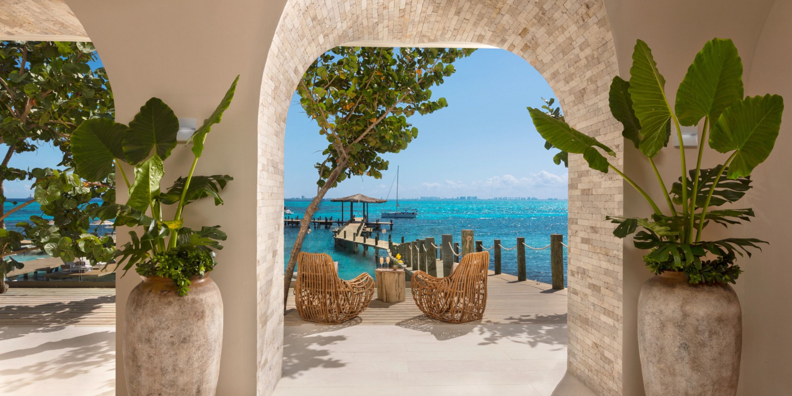 Observe the scenic view from Impression Isla Mujeres by Secrets, featuring two wicker chairs facing a wooden pier and the turquoise ocean. Large potted plants adorn the entrance under a clear blue sky.
