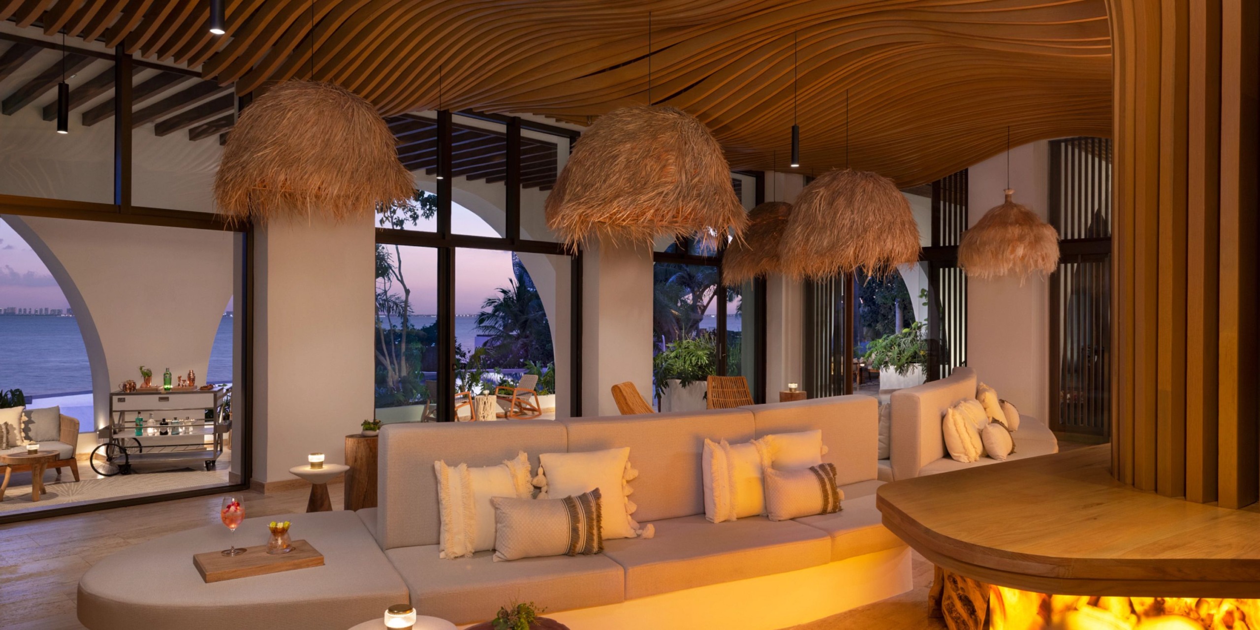 Experience the luxurious lounge area at Impression Isla Mujeres by Secrets, featuring a spacious curved sofa, elegant wooden ceiling, and charming basket-style pendant lights. Glass doors provide an unobstructed ocean view, creating a cozy ambiance with warm lighting.