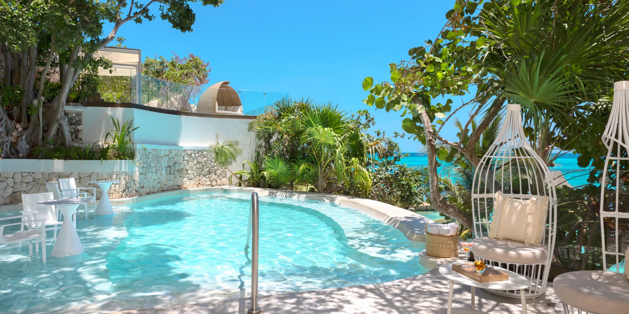 Experience a tranquil poolside ambiance at Impression Isla Mujeres by Secrets, complete with elegant white furniture, lush tropical plants, and breathtaking views of the blue ocean beneath a clear sky.