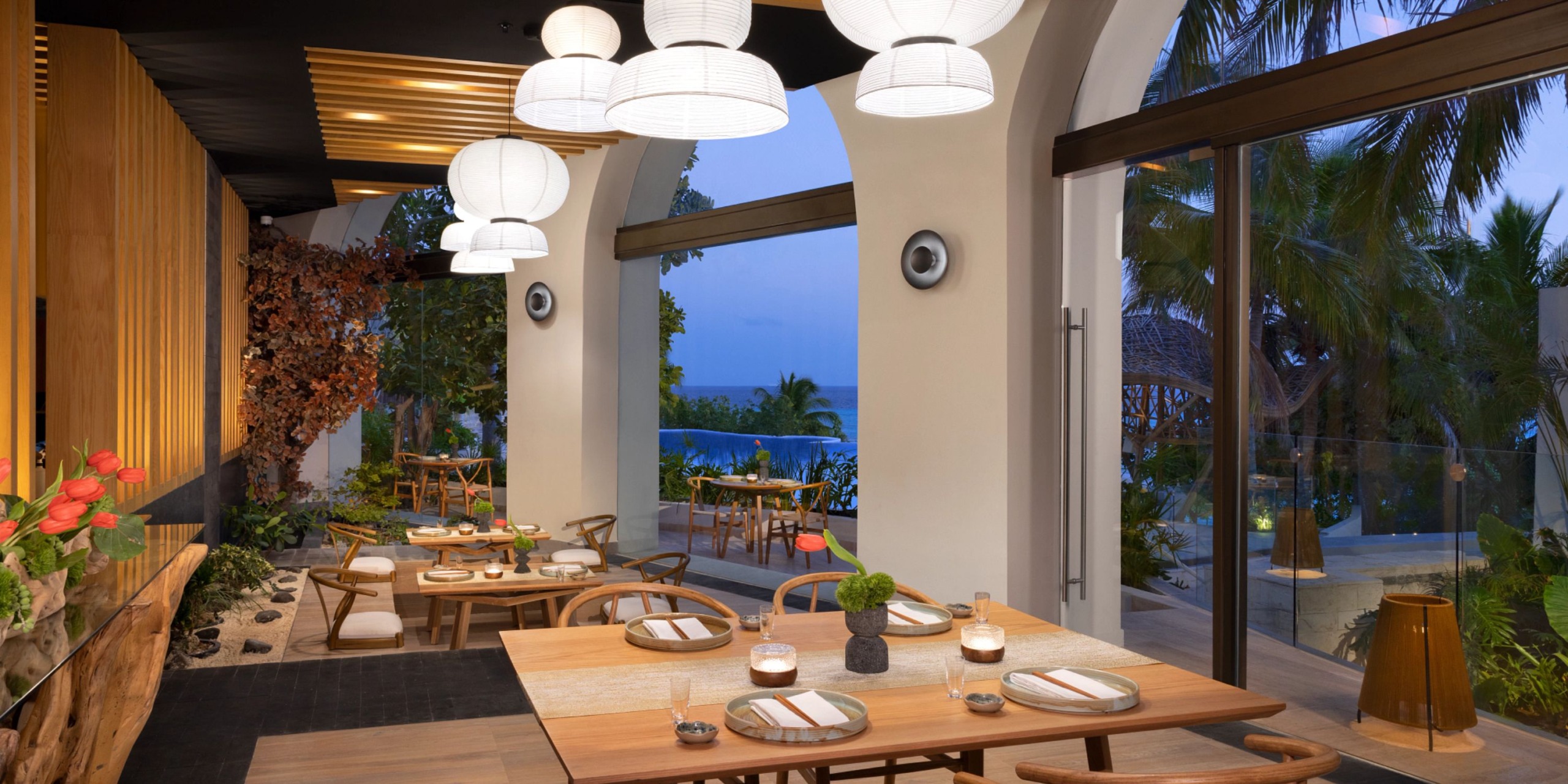 Experience the scenic ocean views at Impression Isla Mujeres by Secrets, an open-air restaurant featuring wooden tables and chairs, pendant lights, and surrounded by lush greenery. Marvel at the breathtaking vista through large windows and arches.