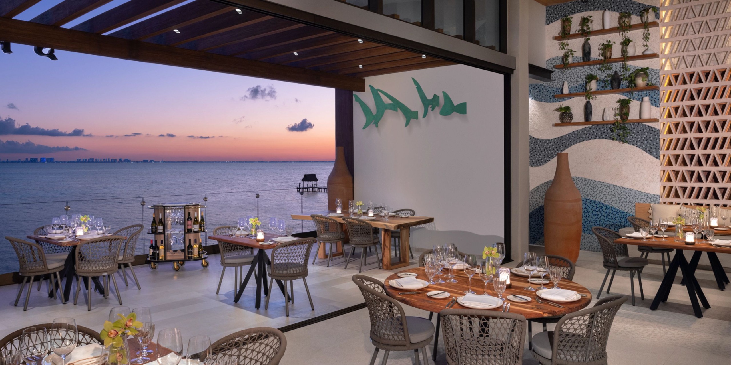 The outdoor dining area at Impression Isla Mujeres by Secrets offers stunning ocean views at sunset, complete with elegant tables set for dinner and decorative wall features.