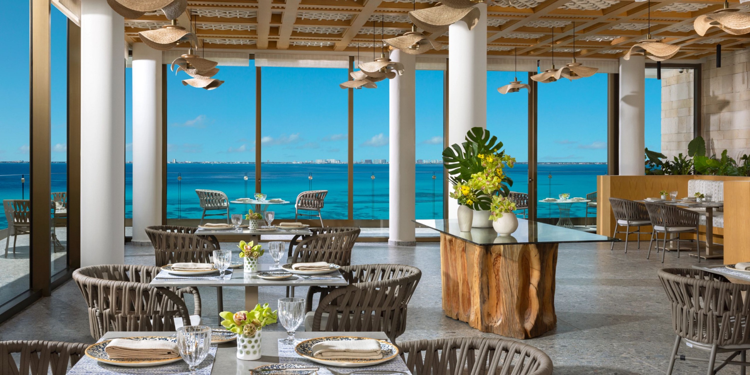 The interior of the Impression Isla Mujeres by Secrets restaurant features woven chairs, set tables, and large windows offering breathtaking views of the clear blue ocean and sky.