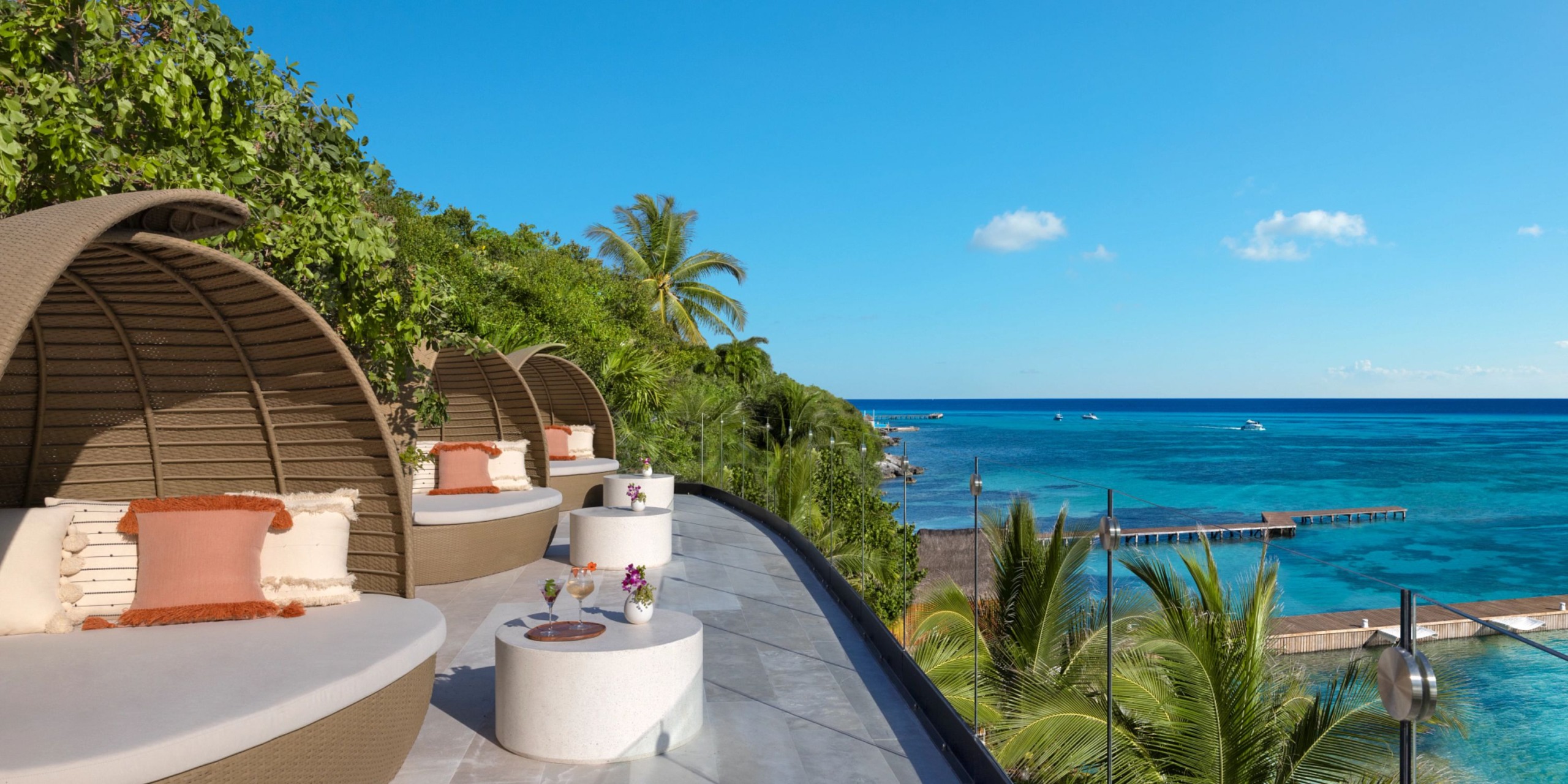 Escape to the Impression Isla Mujeres by Secrets, where you can relax in a modern outdoor lounge area featuring round wicker daybeds adorned with cushions. Enjoy the breathtaking view of a tropical ocean, complete with swaying palm trees and clear blue skies.