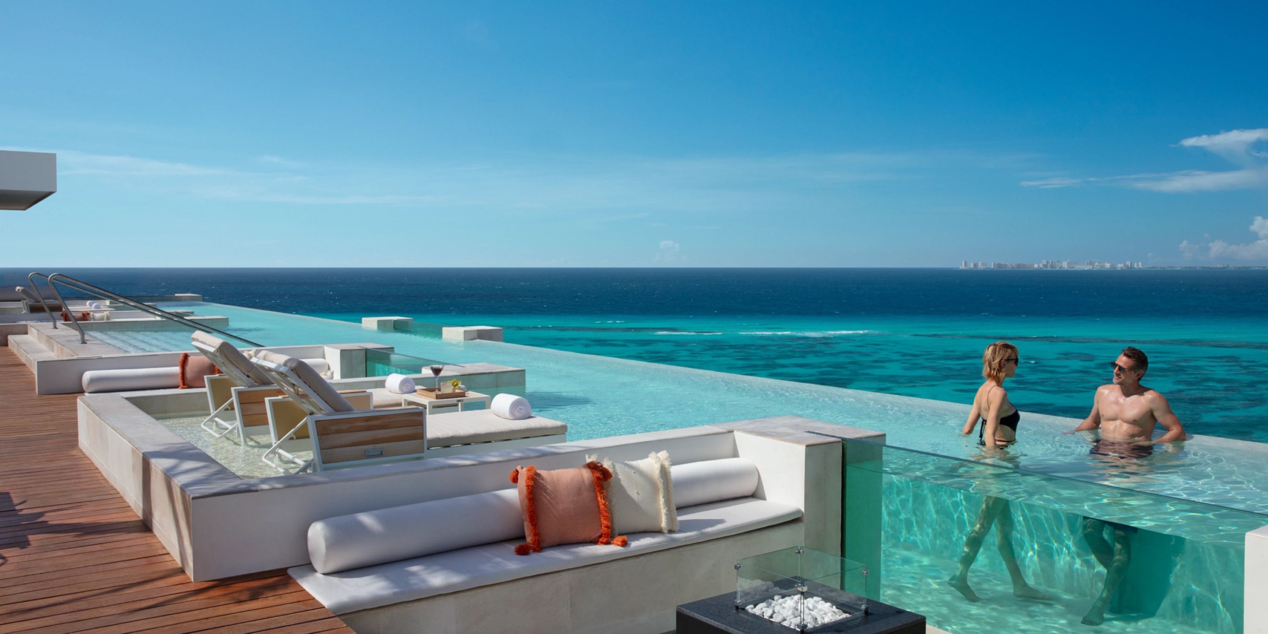 A luxury rooftop infinity pool at the Impression Isla Mujeres by Secrets overlooks the ocean, featuring a couple standing in the water and lounge chairs on the deck.