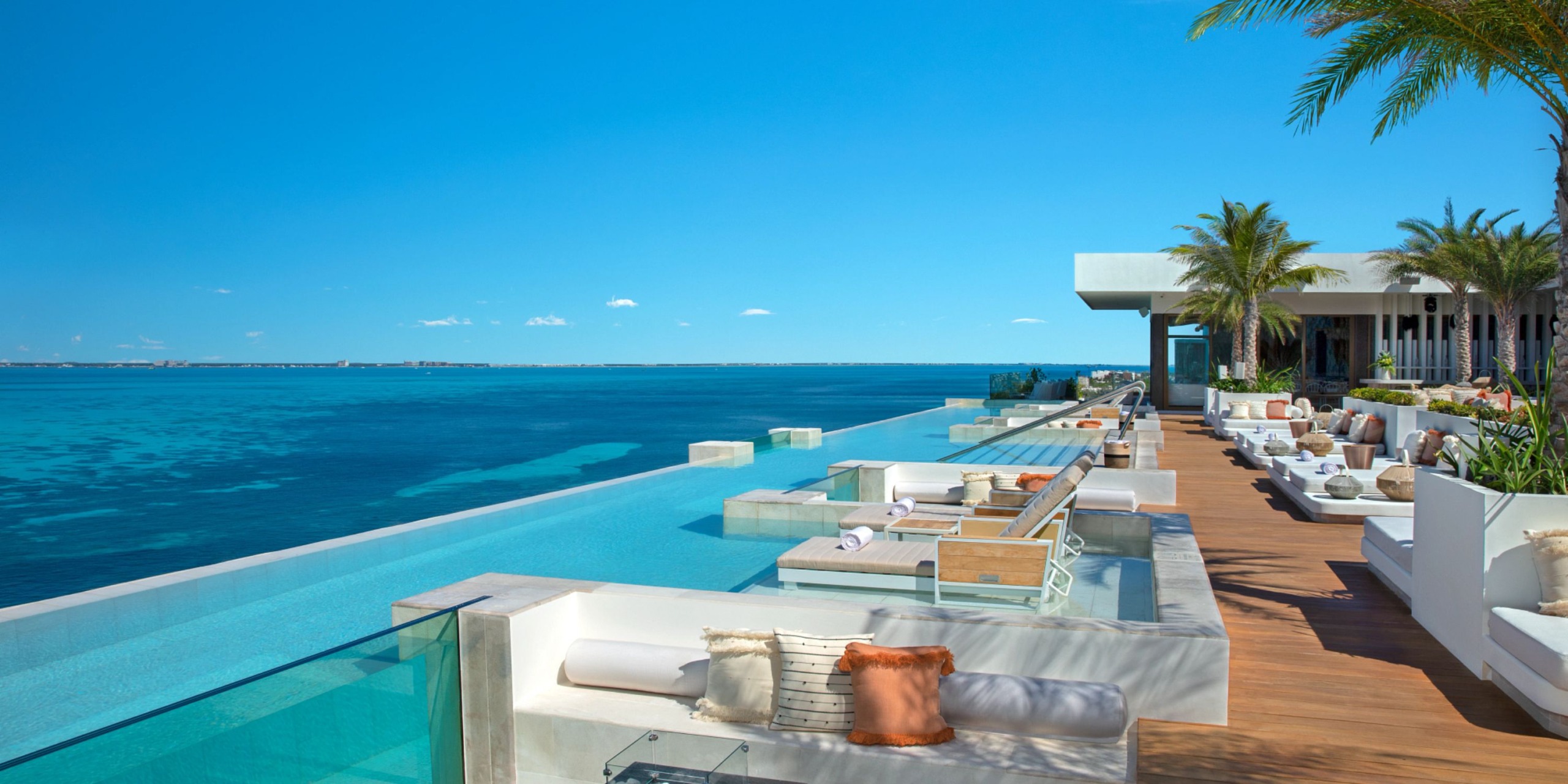 The Impression Isla Mujeres by Secrets offers a modern oceanfront terrace with an infinity pool, comfortable lounge seating, and palm trees, all overlooking a vast blue sea under a clear sky.