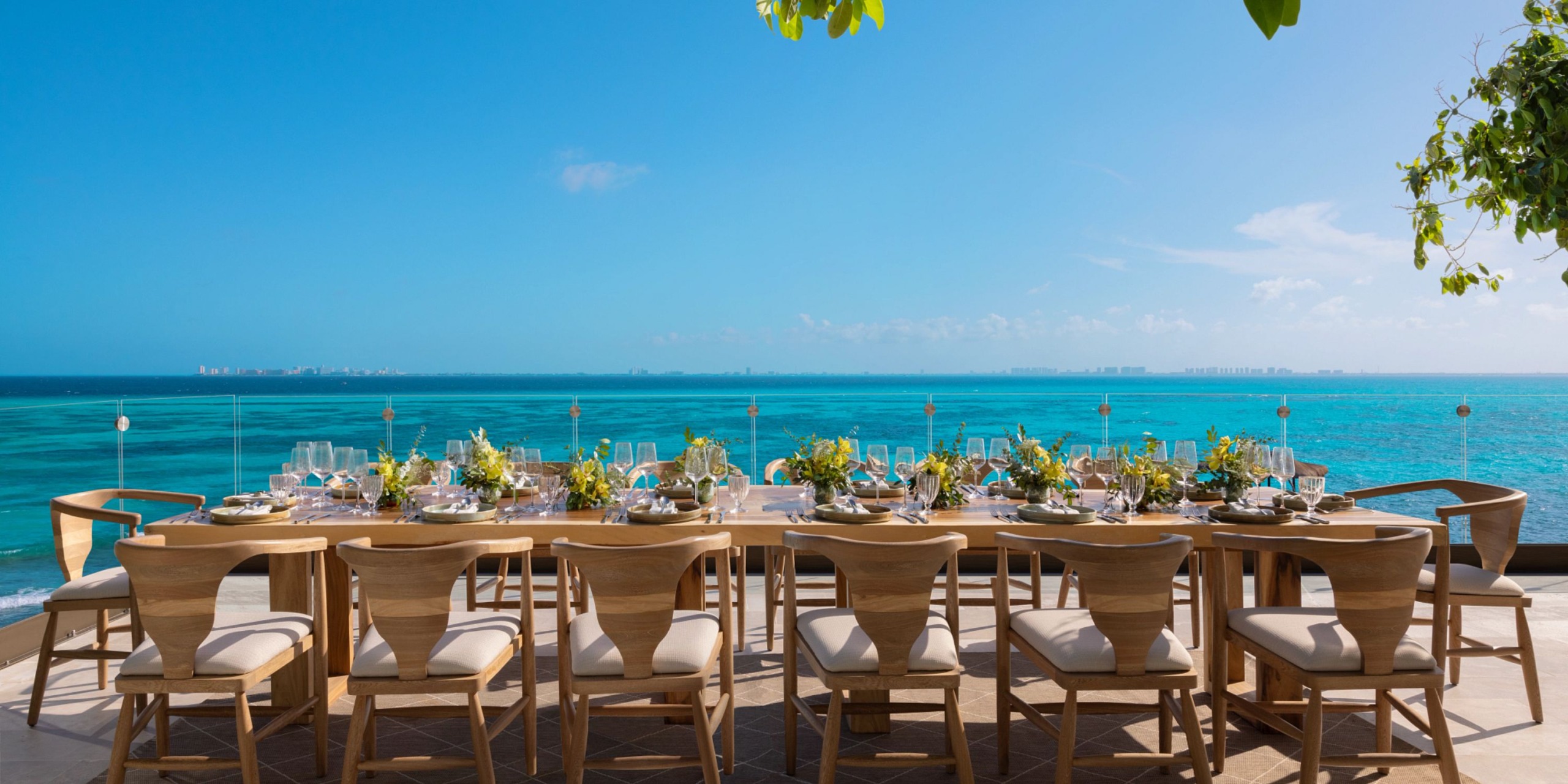 The Impression Isla Mujeres by Secrets dining table set, adorned with floral arrangements, offers a picturesque view of the blue ocean under a clear sky.