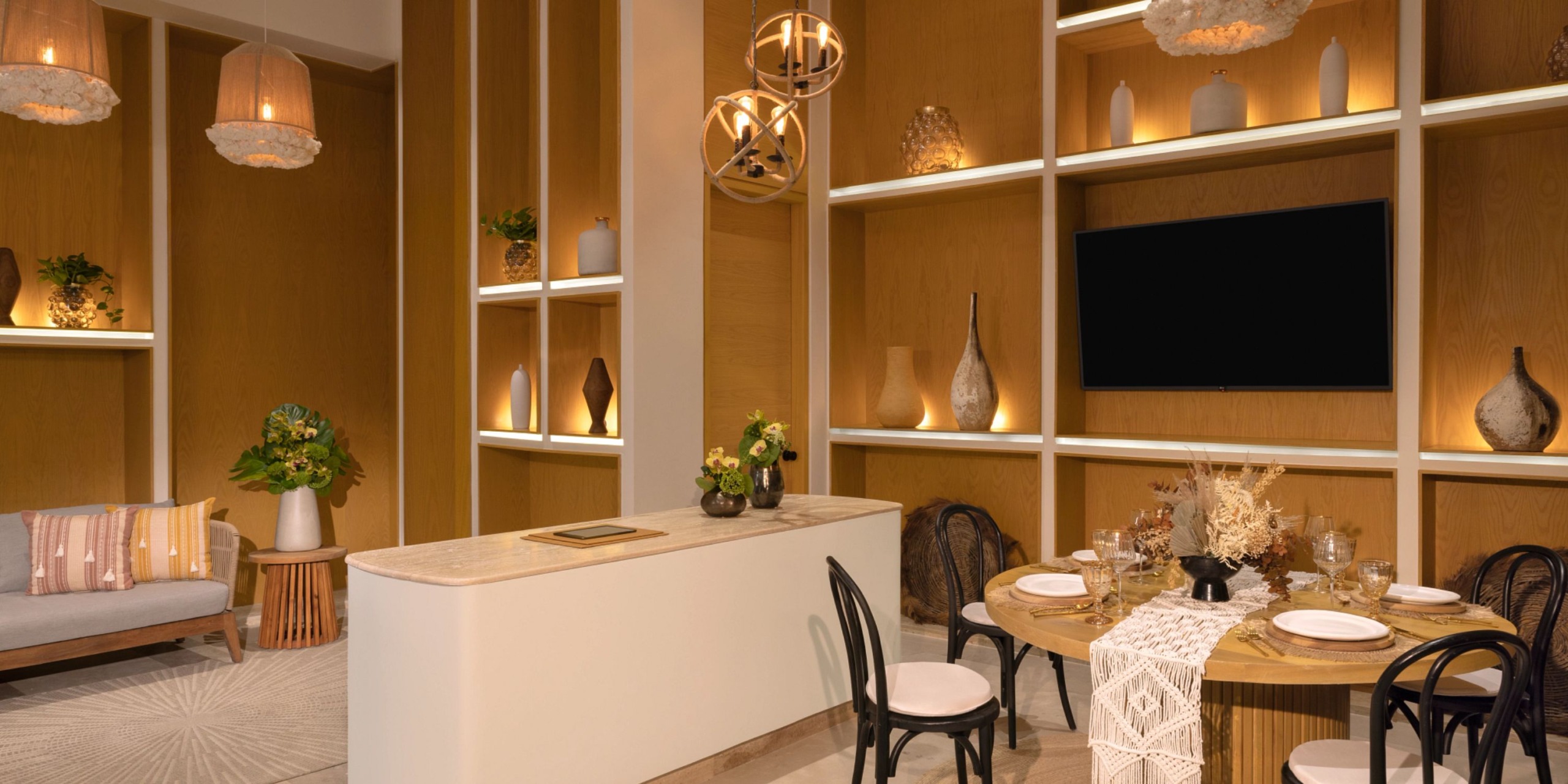 A modern dining area featuring the Impression Isla Mujeres by Secrets with a round table set for four, complemented by beige and brown decor, a wall-mounted TV, and shelves displaying decorative items.