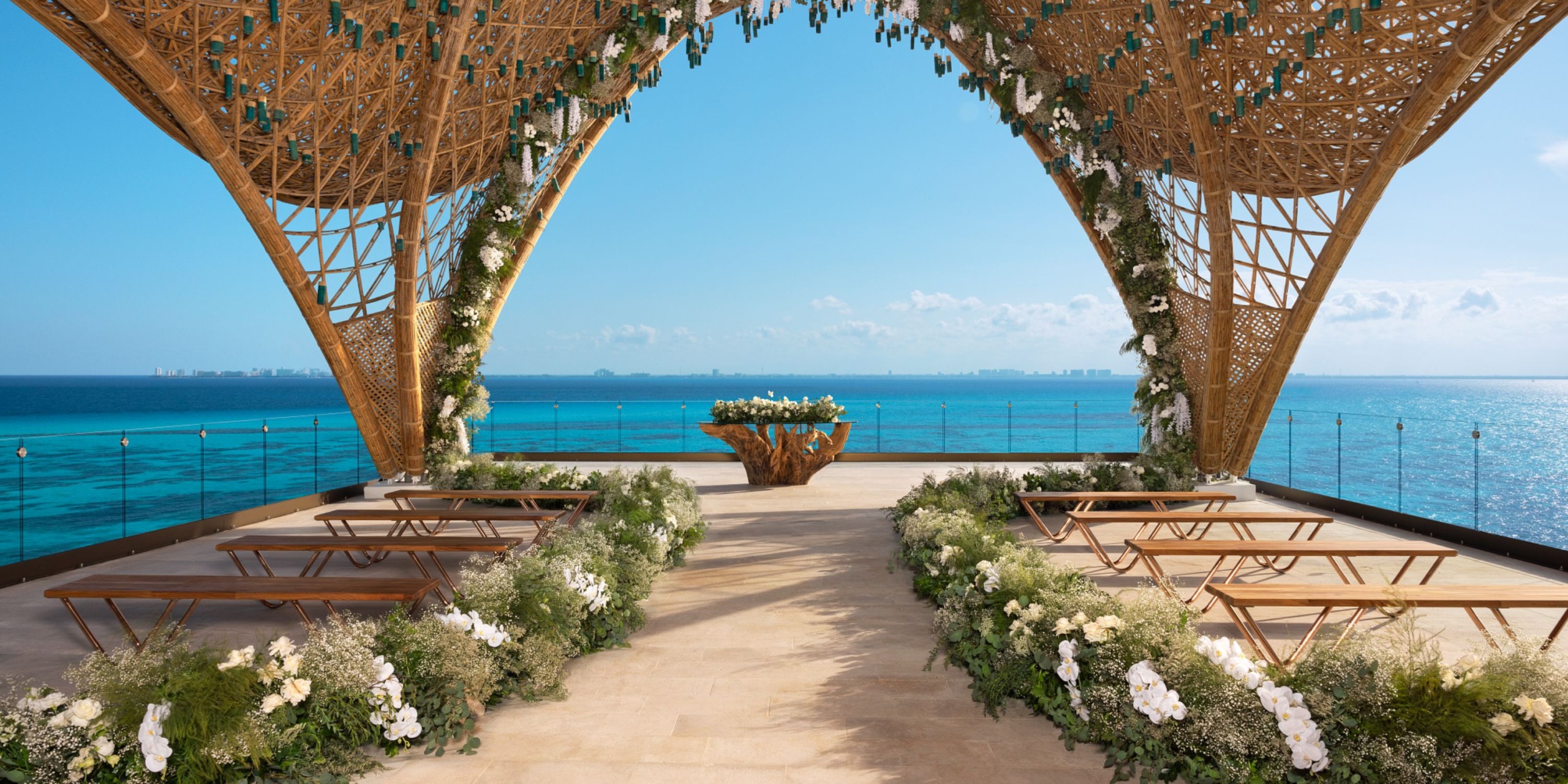 The Impression Isla Mujeres by Secrets features a wooden arch embellished with decorative greenery that provides a stunning view of the ocean. A row of benches is arranged on a stone platform, all directed toward a table beautifully adorned with flowers.
