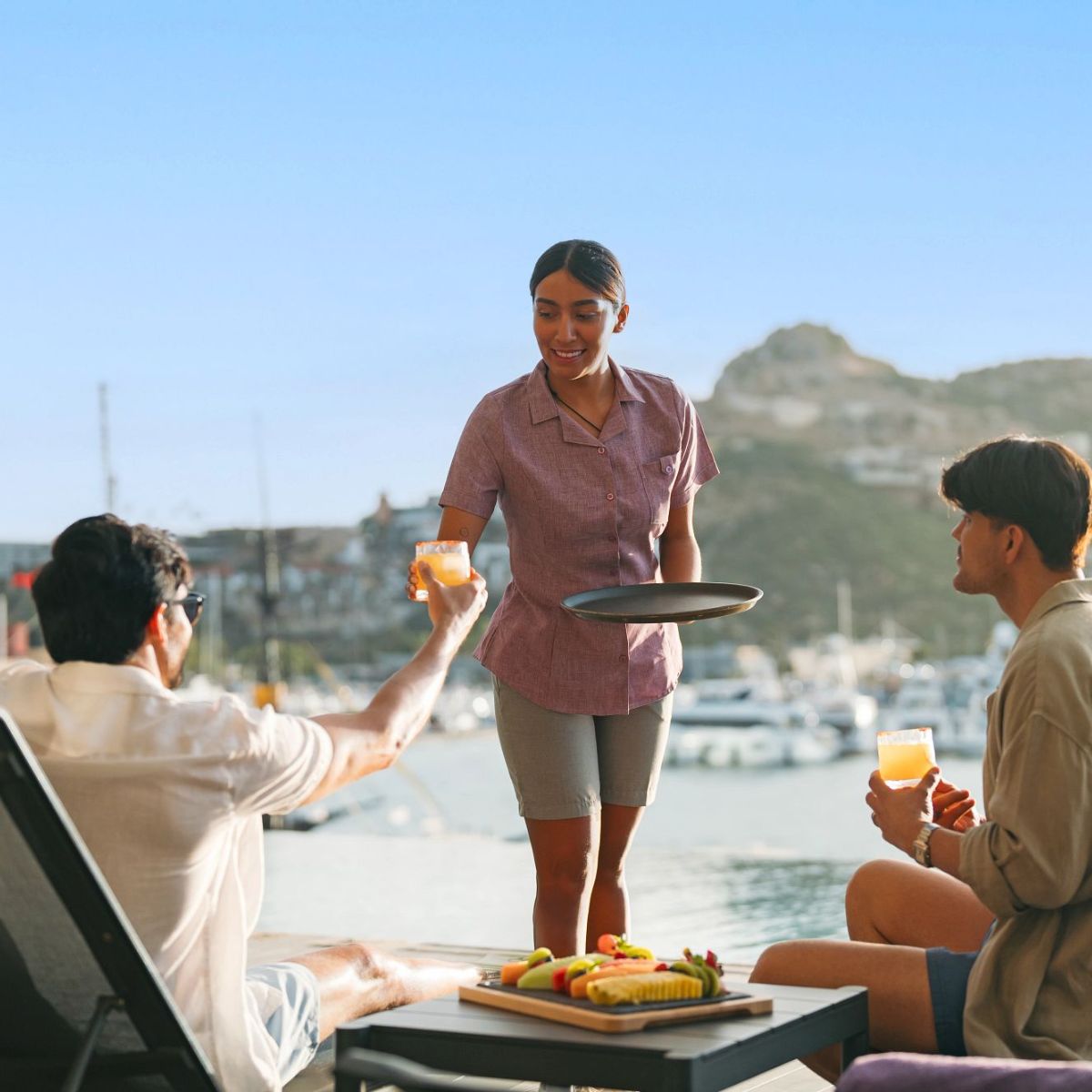 A server hands drinks to two guests by a marina—a beautiful setting for learning proper wedding or destination wedding tipping etiquette.