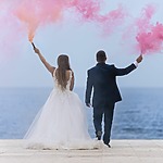 A bride and groom holding pink smoke bombs by the ocean, showcasing their wedding colors.