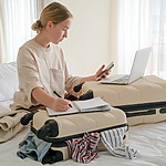 A woman sitting on a bed with luggage and a laptop, preparing for her destination wedding.
