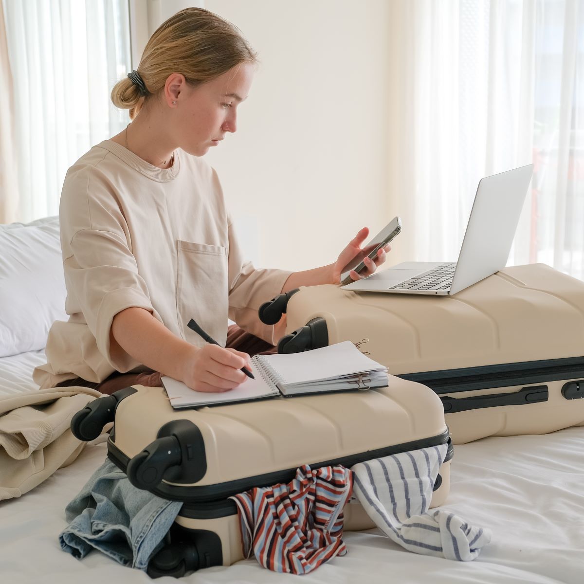 A woman sitting on a bed with luggage and a laptop, preparing for her destination wedding.
