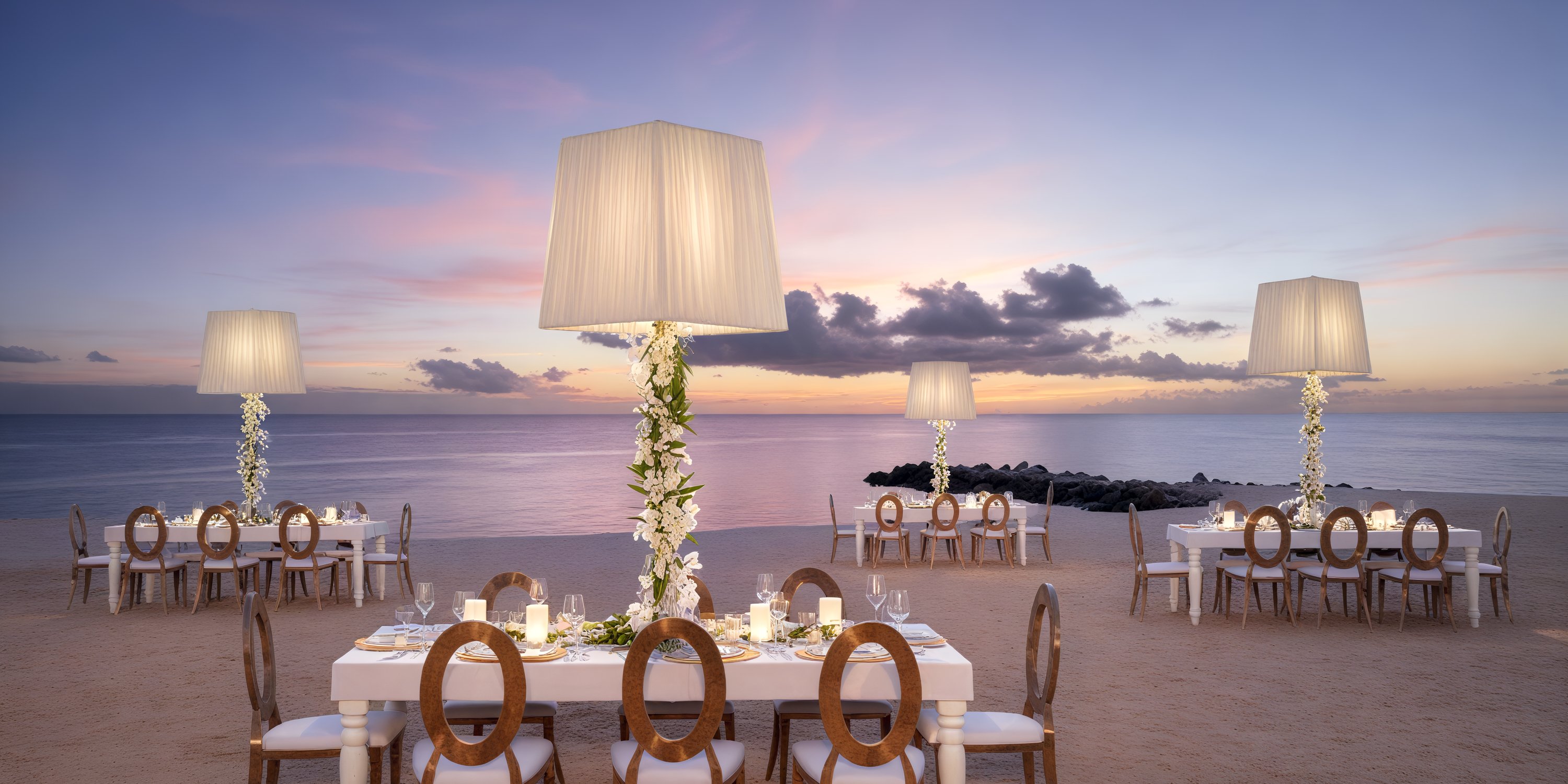 A stunning beachfront dining arrangement featuring tables, chairs, and tall lamp decor at sunset, perfect for a dream destination wedding.