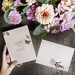 A hand holding a save the date card and flowers on a rustic wooden table for a destination wedding.