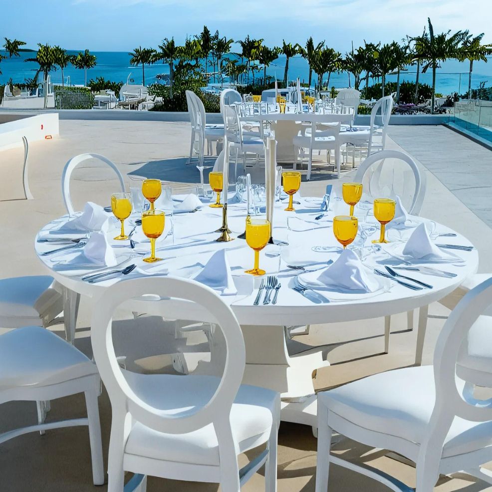 A white outdoor table set with multiple yellow-tinted glasses, white napkins, and cutlery overlooking a scenic view of the ocean and palm trees.