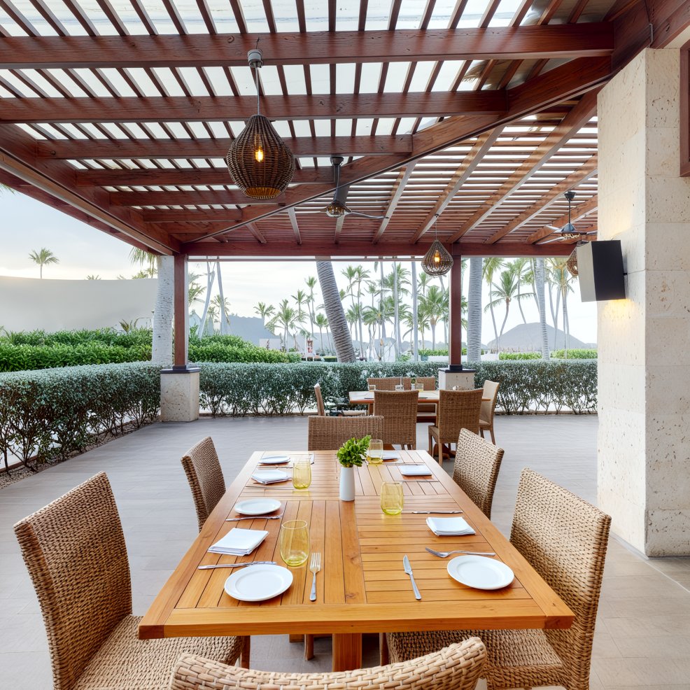 An outdoor patio dining area designed for a destination wedding, featuring a wooden table set with wicker chairs, all overlooking a view of picturesque palm trees.